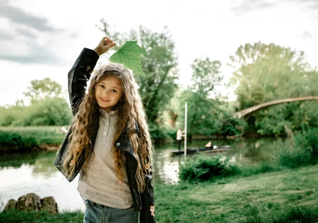 Girl holding a leaf above her head with a punt on the river behind her, Oxford children photographer at University Parks