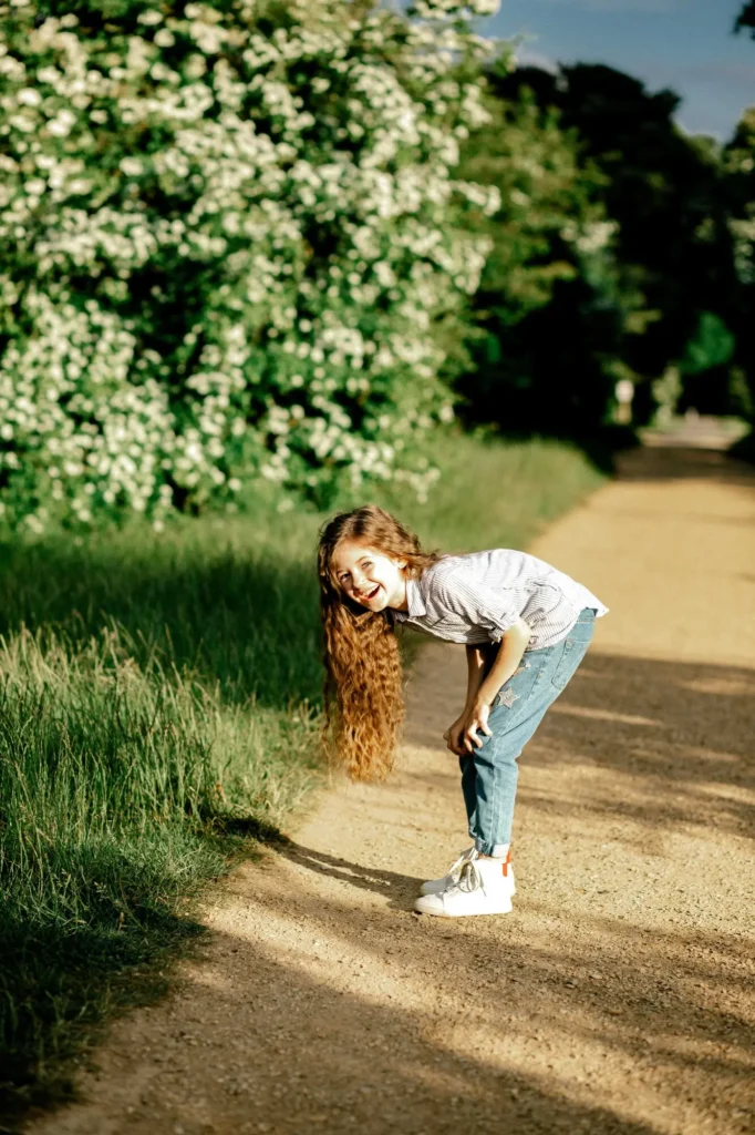 Girl bent over laughing beside a flowering hedge, Oxford children photography at University Parks