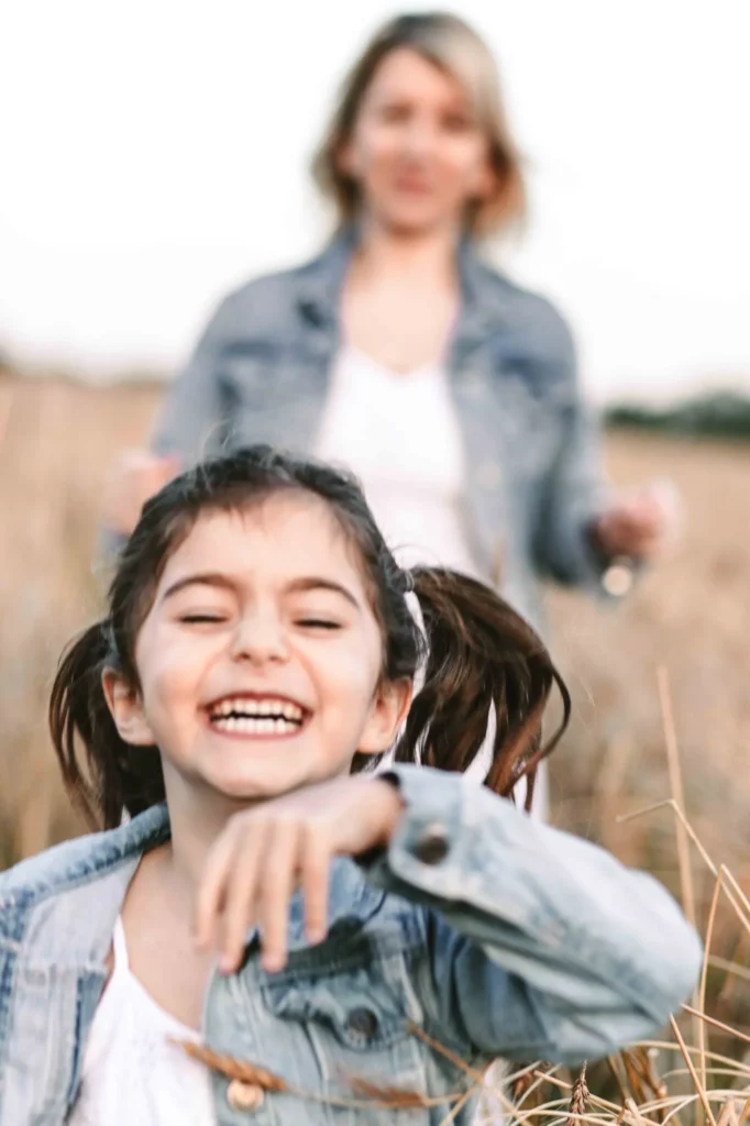 Young girl laughing in a wheat field with her mother blurred in the background, Oxford family photographer